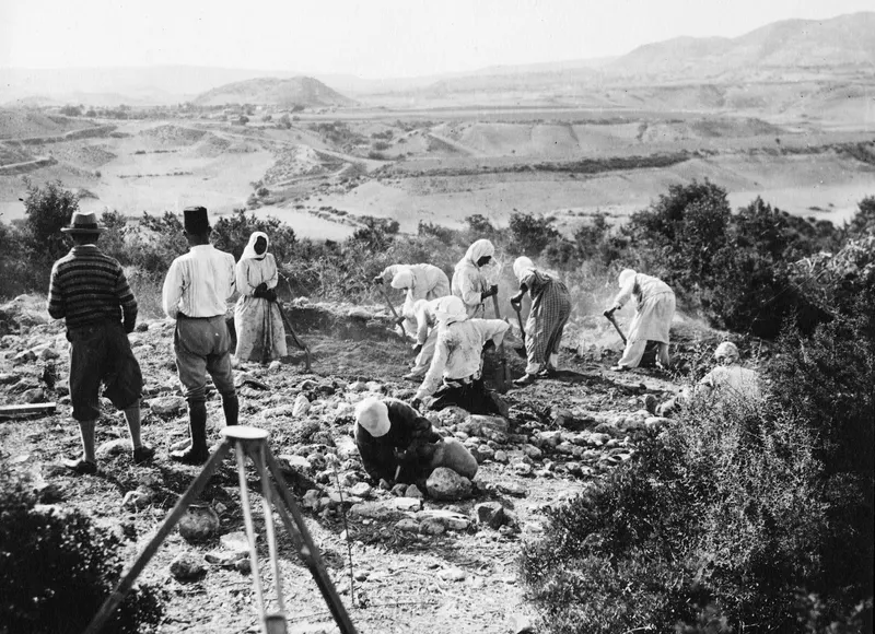 Workers digging with hoes and picks at Palaeoskoutella on a rocky hillside, with a tripod in the foreground and a valley beyond.