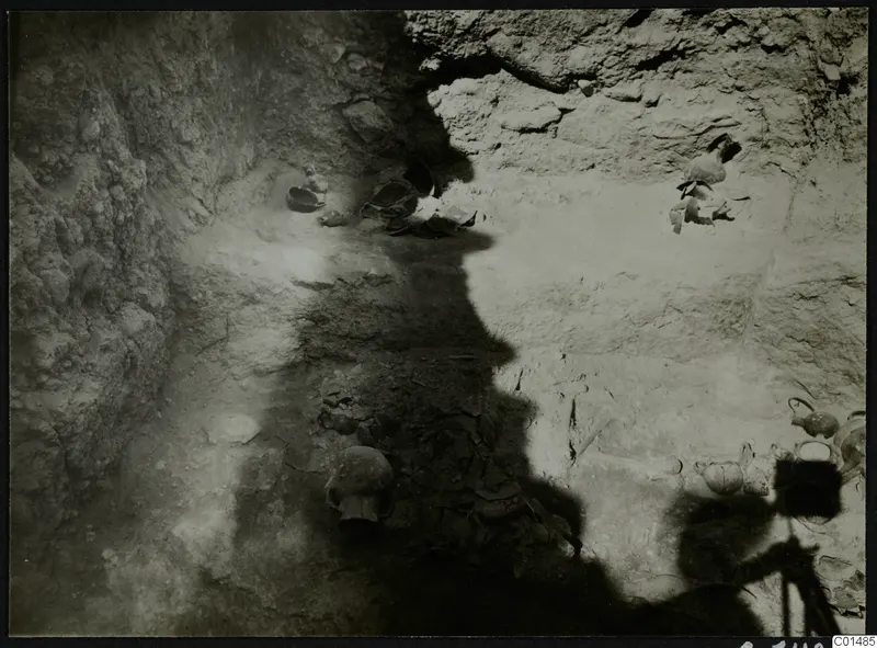 Excavated pit in pale earth with a human skull, broken sherds and several vessels in situ, with a camera tripod at lower right.
