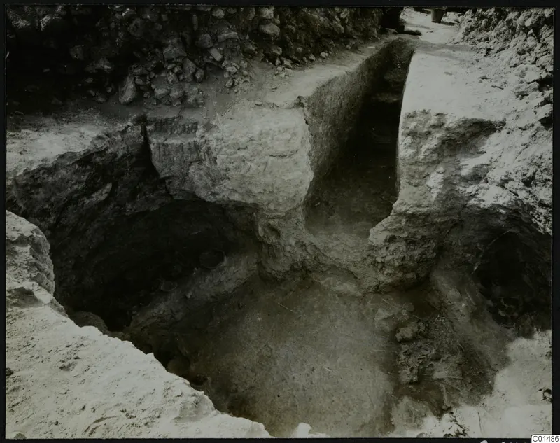 Excavation at Nitovikla showing cut soil faces, a stone wall at the top and small round objects in a dark hollow.