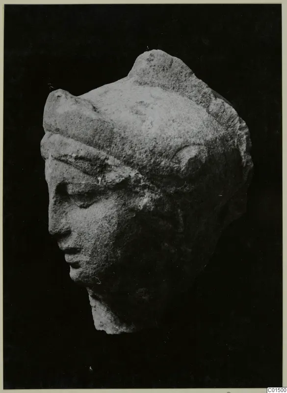 Carved stone head fragment with a band across the brow and downcast eyes, shown against a black background