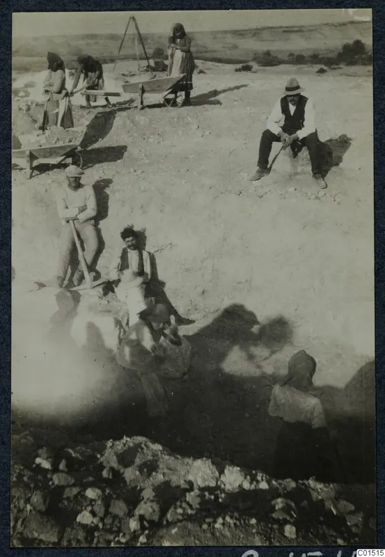 Workers digging at the Soli theatre excavation with picks and a wheelbarrow, with a heap of stones in front