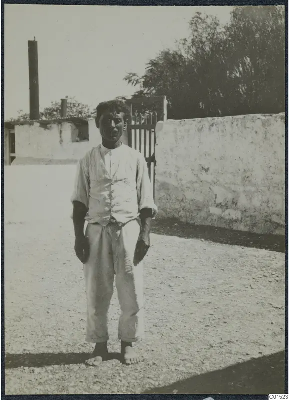 Man in shirtsleeves, waistcoat and long trousers standing barefoot on a gravel yard in front of a whitewashed wall.