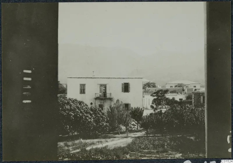 View through a window frame over a whitewashed two-storey house, trees and mountains behind it