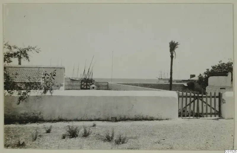 White plastered wall and wooden gate in the foreground, with masted boats and the sea beyond, at Larnaca