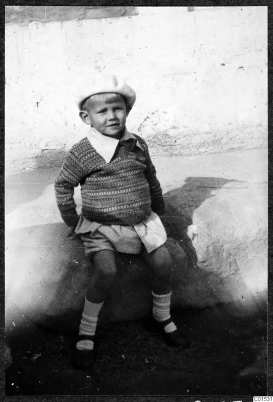 Young child in a knitted jumper and pale beret sitting on a large stone against a whitewashed wall.