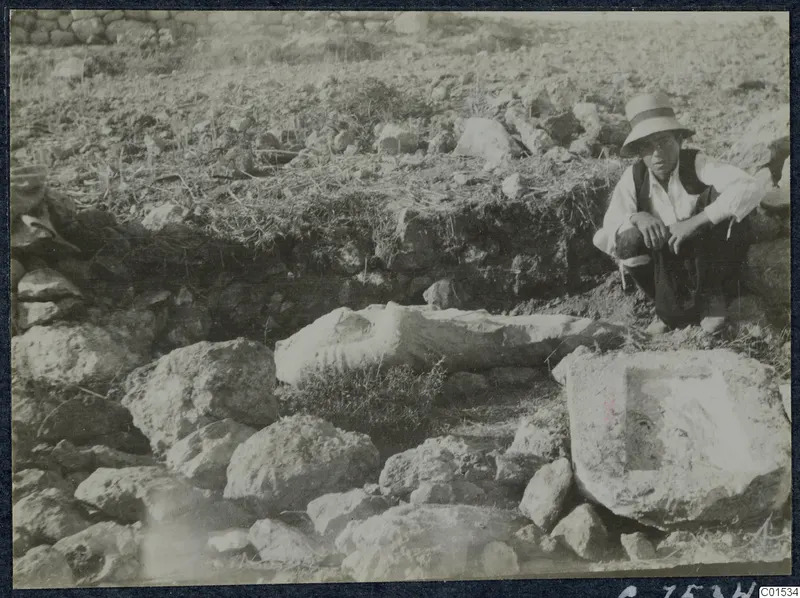 Carved stone fragment and a block with a recess among rubble at Vouni, with a seated man beside them.