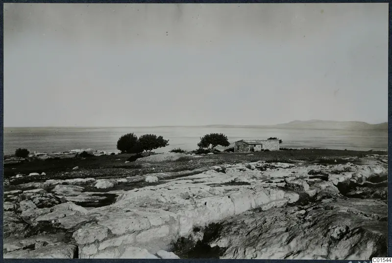 Small stone building and trees on rocky ground at Vouni, with the sea and distant hills behind