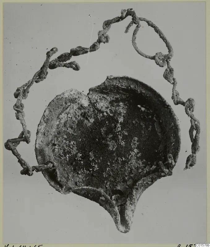 Corroded metal lamp with a shallow round bowl and pointed spout, with a looped metal chain attached
