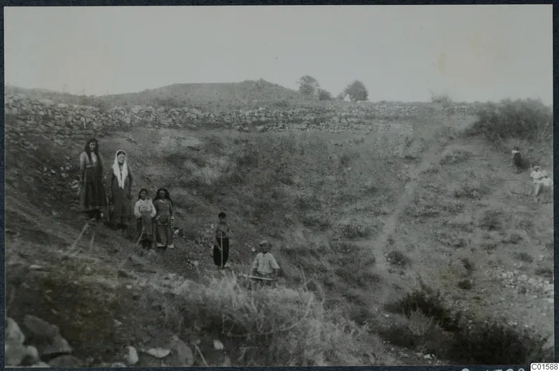 Theatre at Soli before excavation: a grassy slope with a stone wall, and a group of people in the foreground.