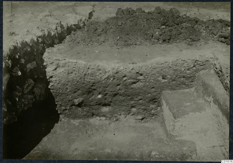 Excavation at the Vouni palace showing a low rubble wall and a stepped stone block beside a trench.