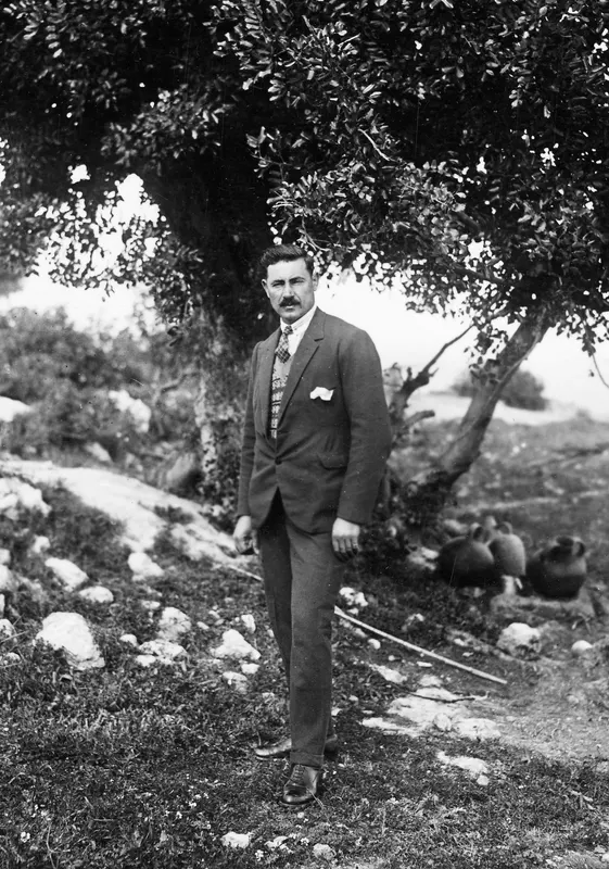 Lazaros in a dark suit standing beneath a large tree at Vouni, with large pottery jars on the ground behind him.