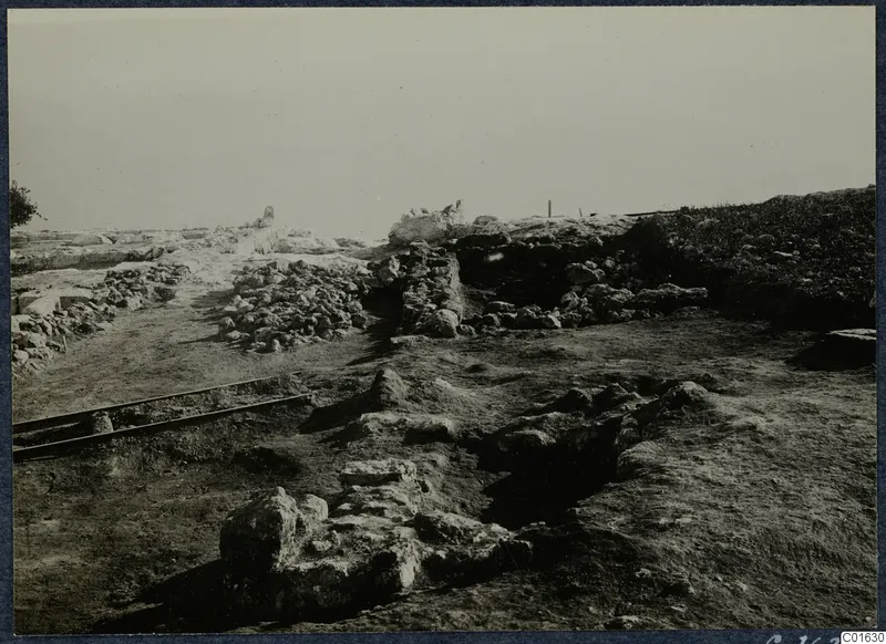 Excavated ruins at the palace at Vouni, with low stone walls, rubble and two rails in the foreground