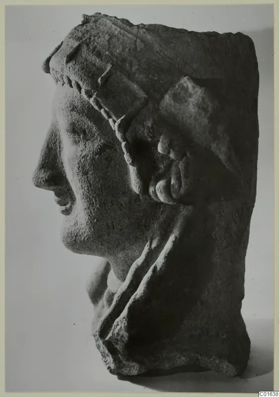 Profile of a carved head facing left, with a beaded band across the brow and a large curled ornament at the ear.