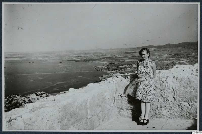 Miss Atterman in a patterned dress standing on a stone wall at Vouni, with the sea and coastal plain behind her.