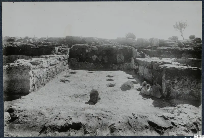 Excavated room at Vouni with stone walls, a pot standing on the floor and a handled jug at the right-hand wall.