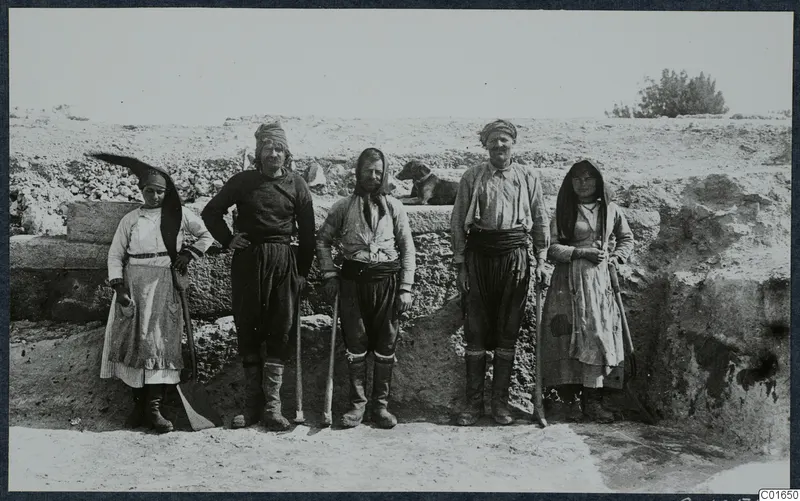 Workers from Tillyria, three men and two women, standing before a stone wall at Vouni, with a dog resting behind them.