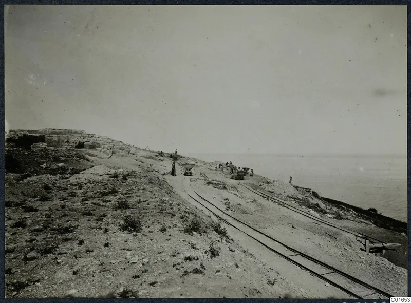 Narrow-gauge rails and small wagons running across a rocky slope above the sea, with cut stone walls at the left and several people.