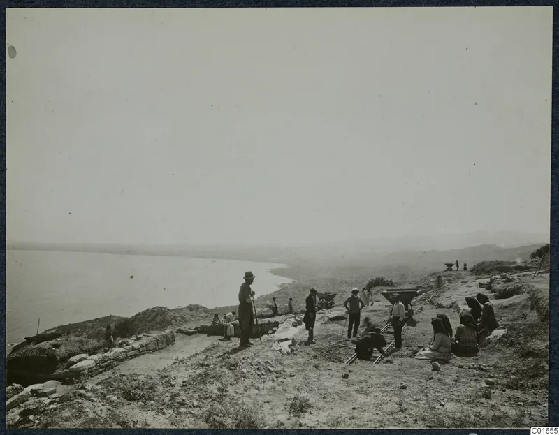 Excavation on a coastal hilltop at Vouni, with workers, a stone wall, rail tracks and the sea to the left.