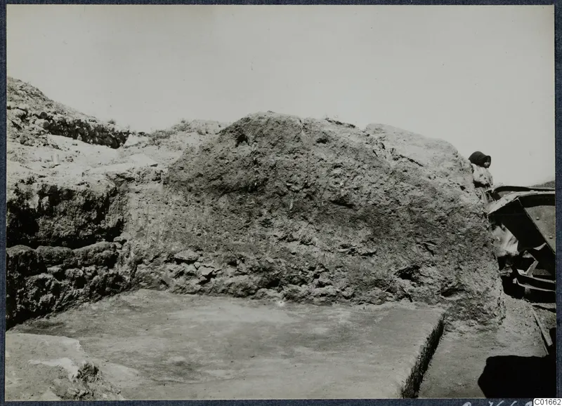 Large earthen wall beside a smooth pale floor at the Vouni palace, with a person and a wooden barrow at the right edge.