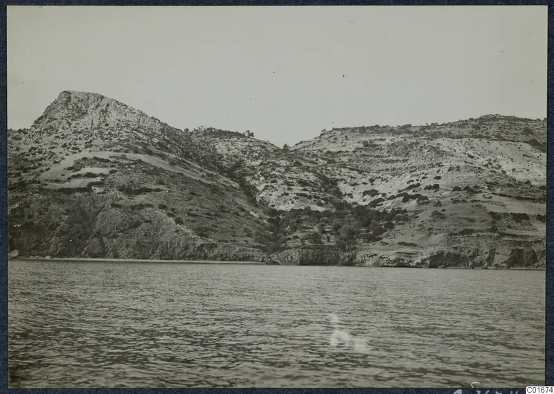 View from the sea of a rocky, scrubby headland rising above a cliff-lined shore at Vouni