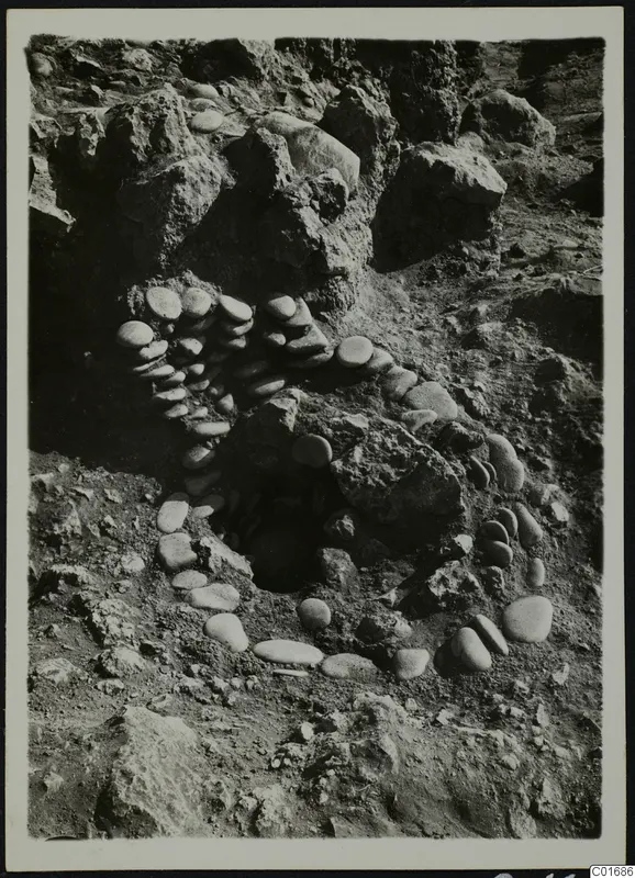 Rounded stones set in a curving line around a dark opening in rocky, excavated ground at Petra tou Limniti