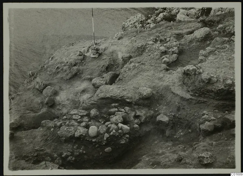 Excavation at Petra tou Limniti with exposed stones and a vertical ranging pole on a sloping earth surface