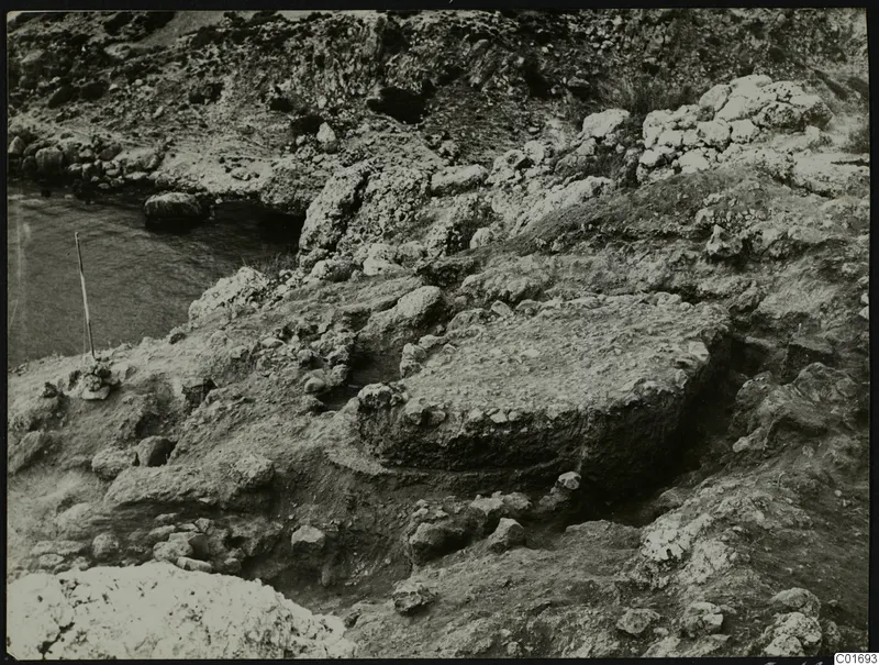 Excavation at Petra tou Limniti with rock ledges, rubble and water at the left edge of the view.