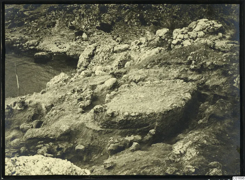 Excavation among rocks at Petra tou Limniti, with water to the left and a thin wooden pole at the edge