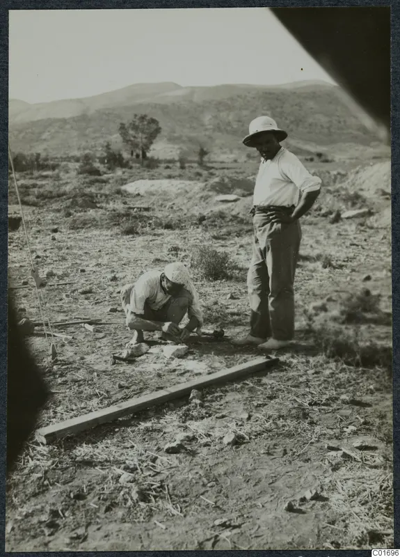 Two men at work on a dry hillside, one crouching and one standing beside a wooden plank on the ground.
