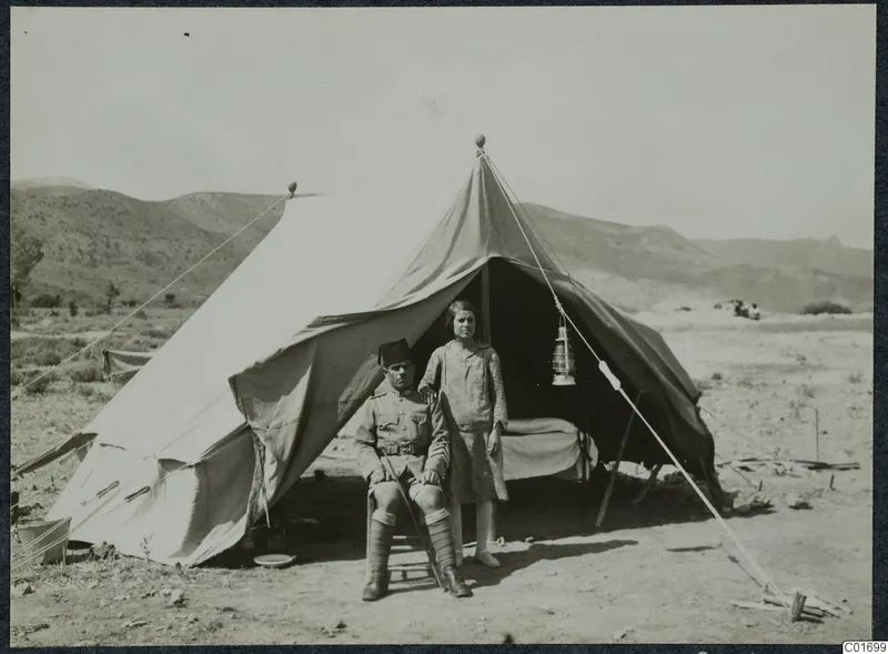 Policeman seated outside a large canvas tent at Ayios Iakovos, with a young girl standing beside him.