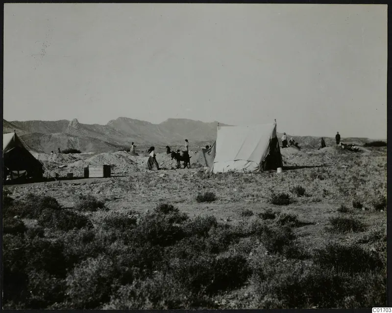 People and white canvas tents on open scrubland at Ayios Iakovos, with mountains behind