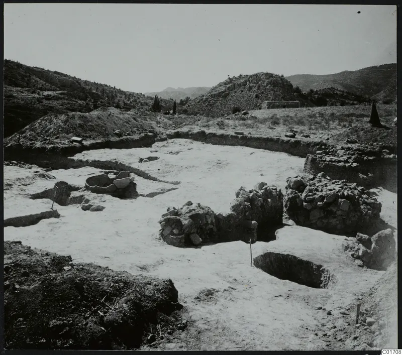 Excavation at Ayios Iakovos seen from the east, with low stone walls and a round stone feature in pale exposed soil.