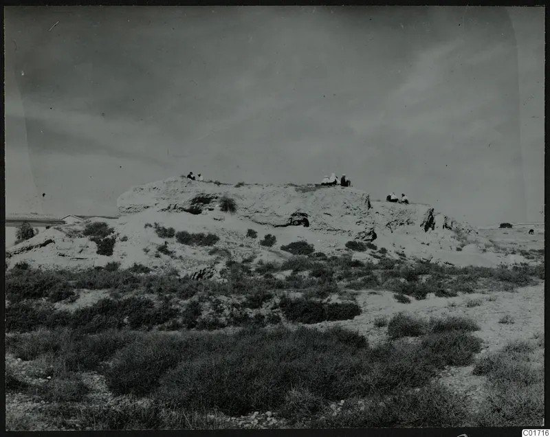 Bamboula mound seen from the southeast, with several people sitting on its top edge above low scrub.