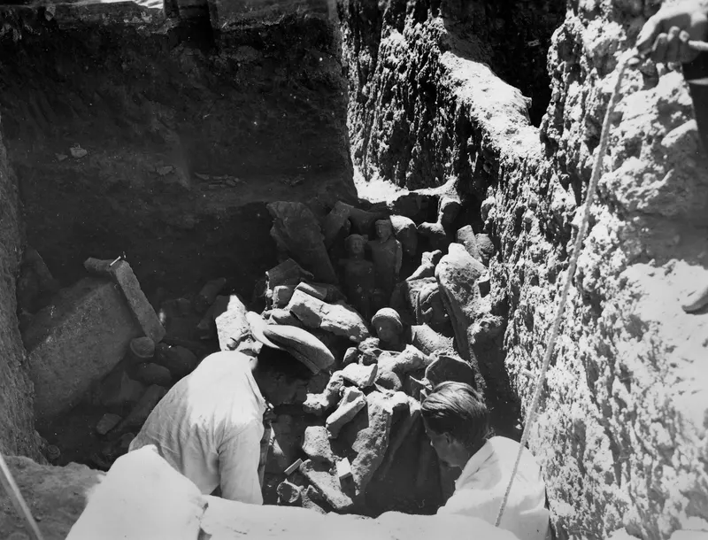 Two men digging among broken stone sculptures in a deep excavation trench at Kition