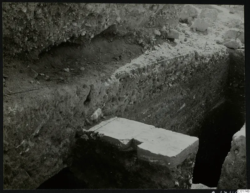 Deep excavation trench with a tall soil section and a large pale stone block at its base, Kition