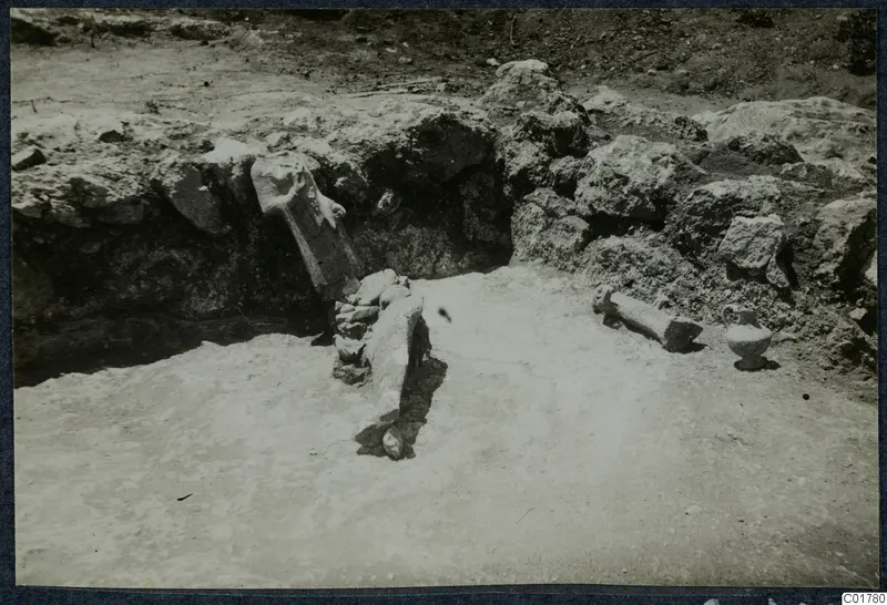 Finds in situ during excavation at Vouni: a small pot and carved stone fragments on a sandy floor below a rubble wall.