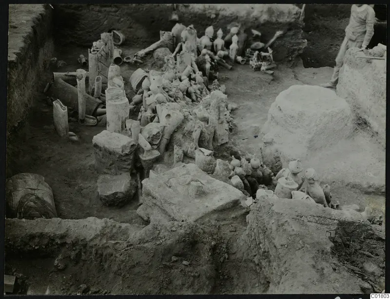 In situ figurines and vessels lying in an excavation trench at Ayia Irini, with a person standing at the edge