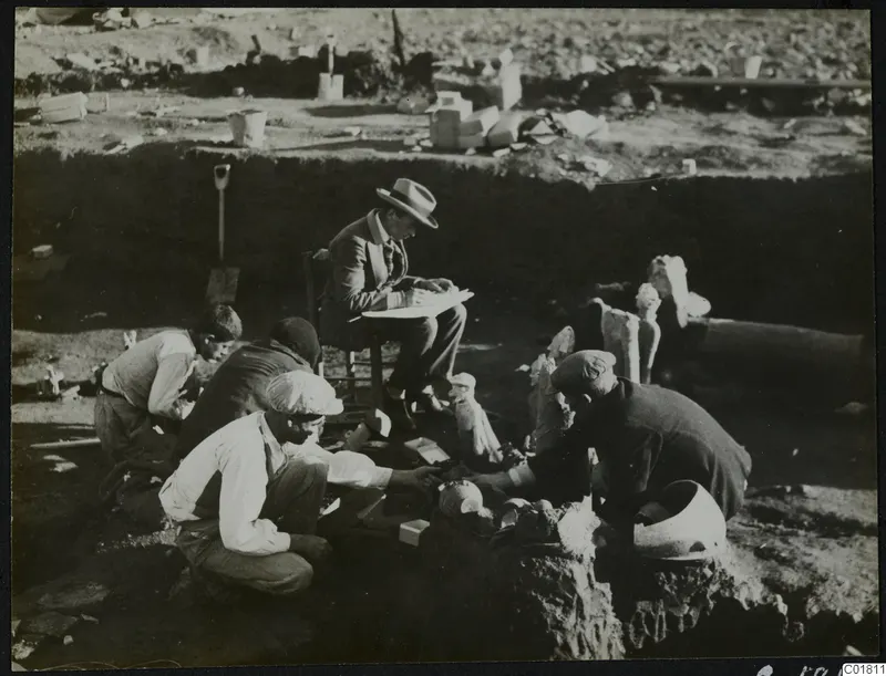 Excavation at Ayia Irini: a seated man in a hat writes while four workers clean and sort broken finds on the ground