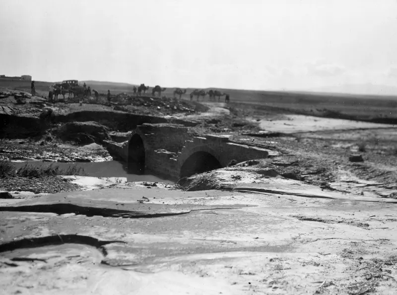 Stone arched bridge over a muddy stream at Chiloura, with a camel caravan and a vehicle on the ridge behind.