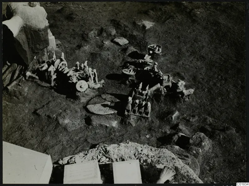 Small pale figurines and wheeled stands lying in the ground during excavation at Ayia Irini.