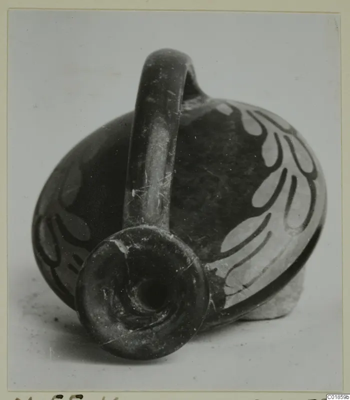 Dark pottery vessel with a strap handle and pale painted leaf motifs, shown from the spout end