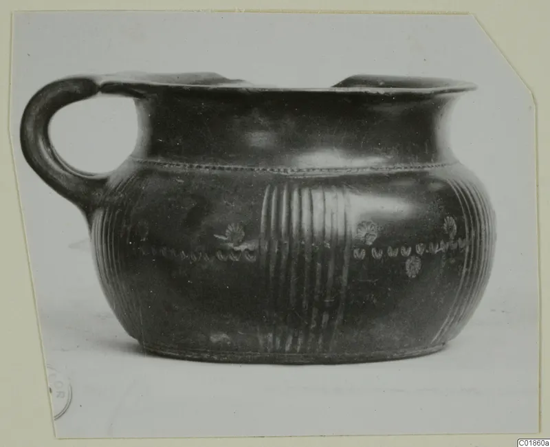 Black-glazed pottery vessel with a loop handle, reeded bands and stamped rosettes, photographed from the side