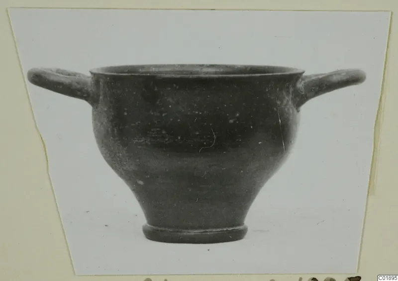 Black glazed skyphos with two horizontal handles, photographed against a pale background on a card mount