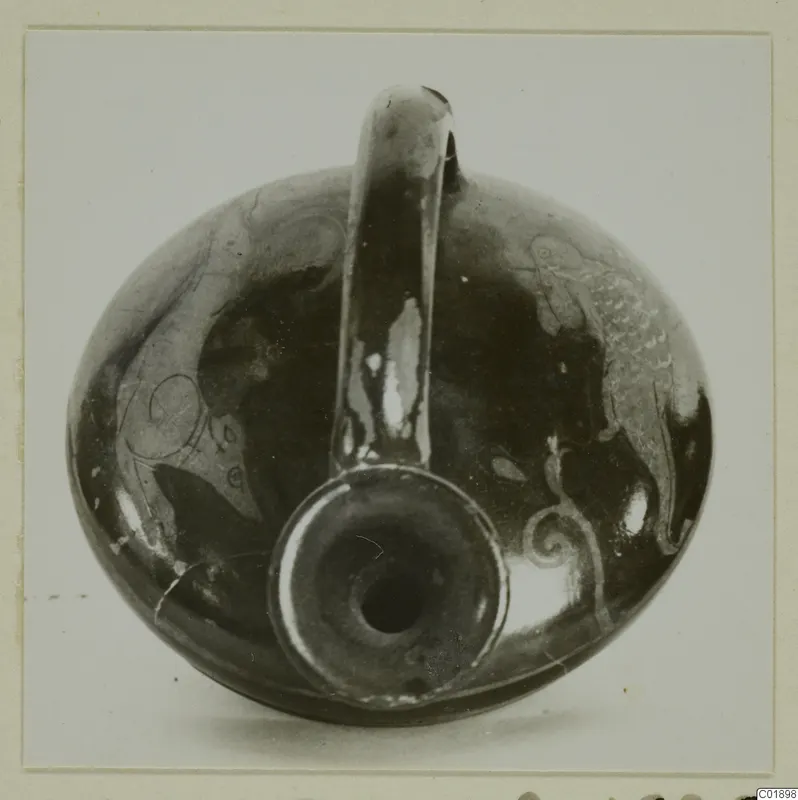 Dark glossy pottery vessel with a tall loop handle and pale painted motifs, photographed from the front.