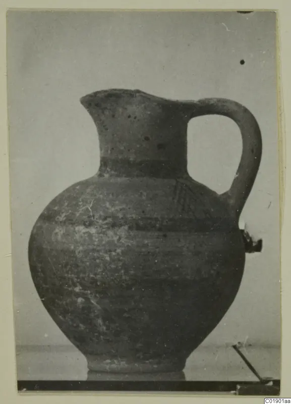 Pottery White Painted I jug with a tall neck, one loop handle and a round body, photographed against a grey backdrop.