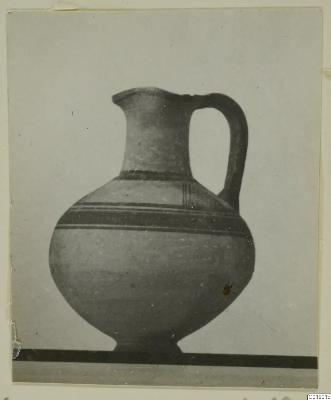 Pottery jug with a high loop handle and dark painted bands on the shoulder and body, shown against a grey backdrop.