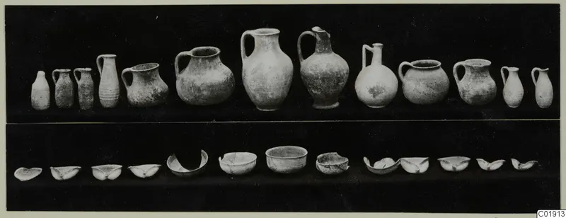 Pottery from Vouni Tomb 13: a row of jugs and juglets above a row of bowls and bowl fragments, on a dark background.