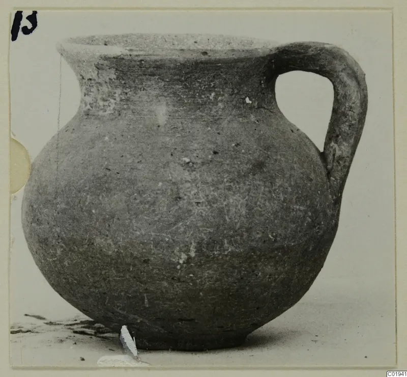 Coarse ware jug with a single handle and a short flared neck, shown in grey-brown clay against a pale background.