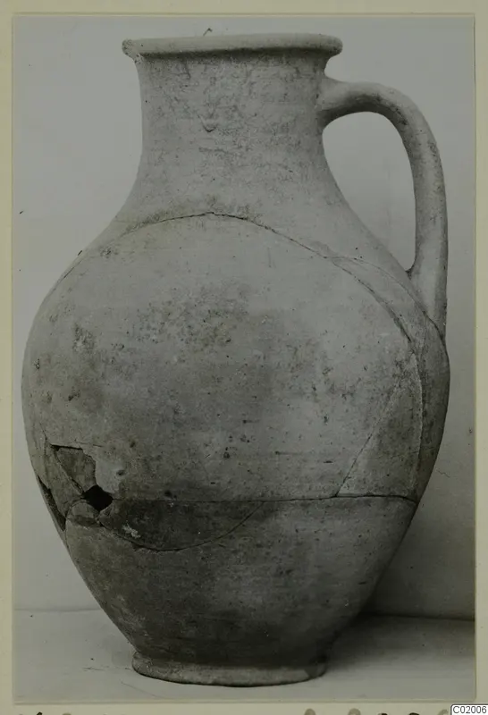 Plain pale pottery jug with a single handle, a flared rim and a cracked, repaired body, shown against a grey backdrop.