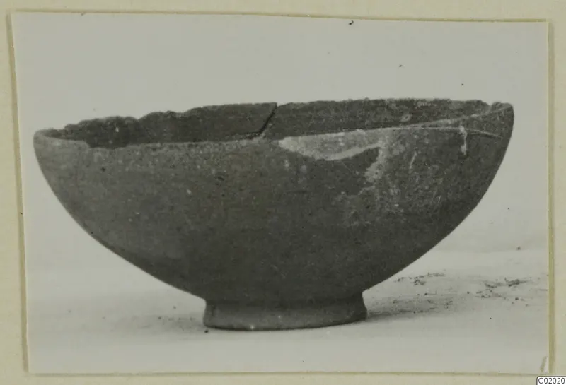 Footed pottery bowl with a broken, uneven rim, shown in side view against a pale background